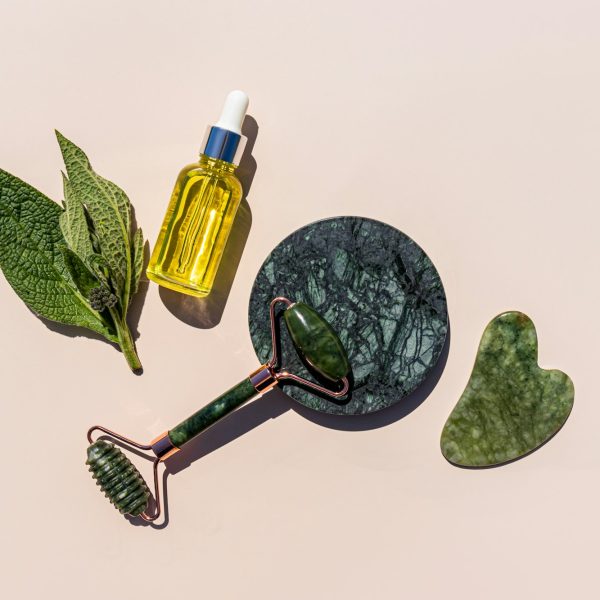 Natural cosmetic massage oil, roller massager and natural jade scraper on a beige background.